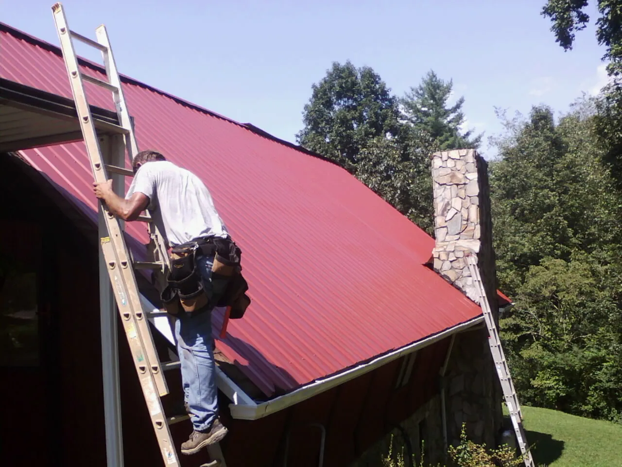 Roofer on a ladder beside a red metal roof and stone chimney — Soffit & Fascia Repair by Roofing Brothers in Johnson